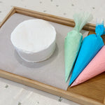 White cake on a wooden tray with colorful piping bags.