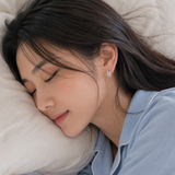 A woman with dark hair sleeping peacefully on a white pillow, wearing light blue pajamas and a small silver geometric huggie earring.