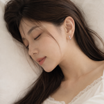 A woman with long dark brown hair rests on a white pillow, wearing a delicate silver crescent-shaped stud earring with a small blue gemstone.