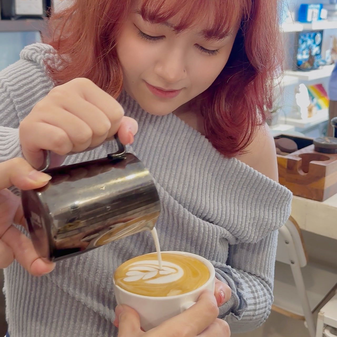 Person pouring milk into a cup of coffee with latte art, in a casual indoor setting.
