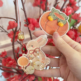A hand holds two decorative paper-cut style ornaments: a peach-shaped ornament with small yellow pom-poms and a bunny ornament with gold and pearl details, in front of a blurred background of red flowers and branches.