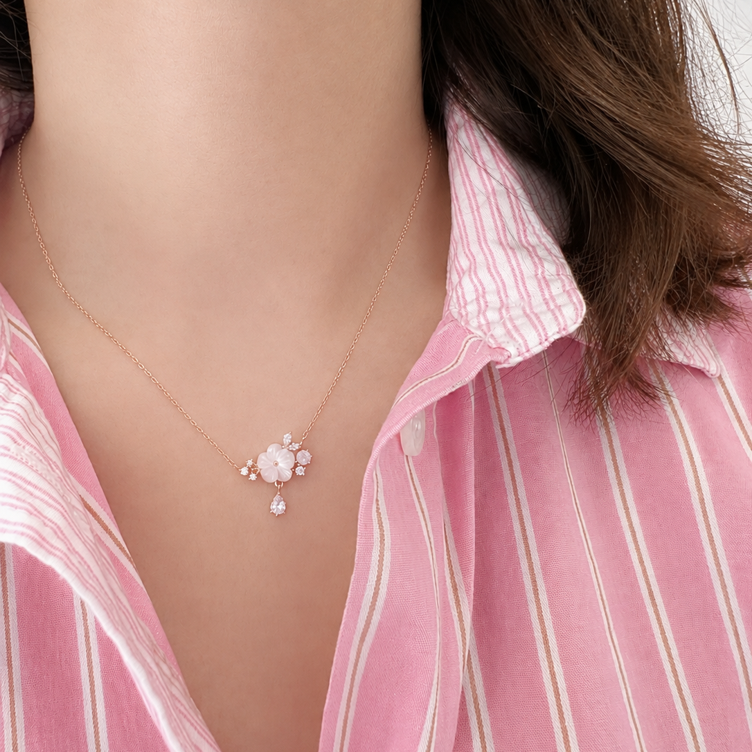 A close-up of a woman wearing a delicate gold necklace with a white floral cluster and a small teardrop pearl pendant, paired with a pink and white striped shirt.