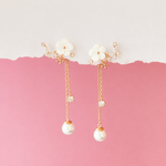 A pair of gold-tone dangle earrings with white flower charms, small clear crystal accents, and white pearl drops, displayed against a pink and white split background.