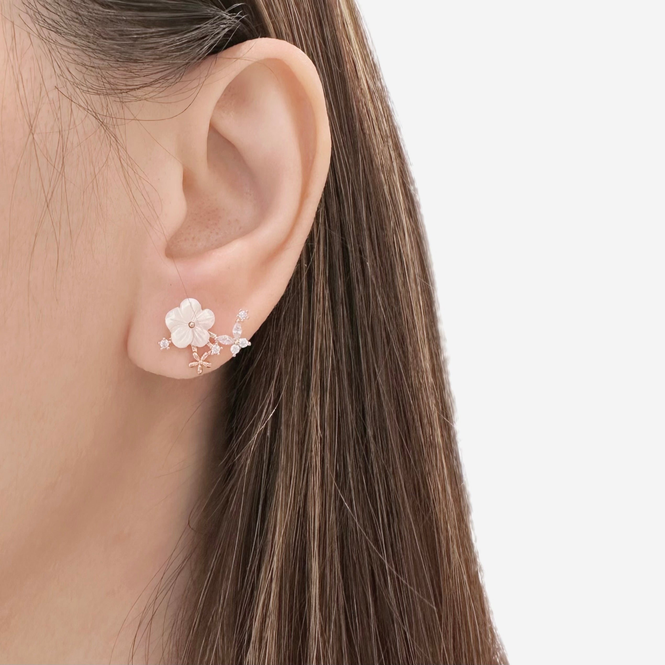 A close-up of a model's ear wearing a delicate floral ear cuff earring in rose gold with white flower petals and small clear stones, set against a clean white background.