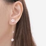 A close-up of a model's ear wearing a rose gold ear cuff with white flower and pearl details, set against a clean white background.