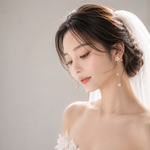 A close-up portrait of a woman wearing a white lace bridal dress and veil, with matching dangling pearl-like earrings, against a soft light gray studio background.