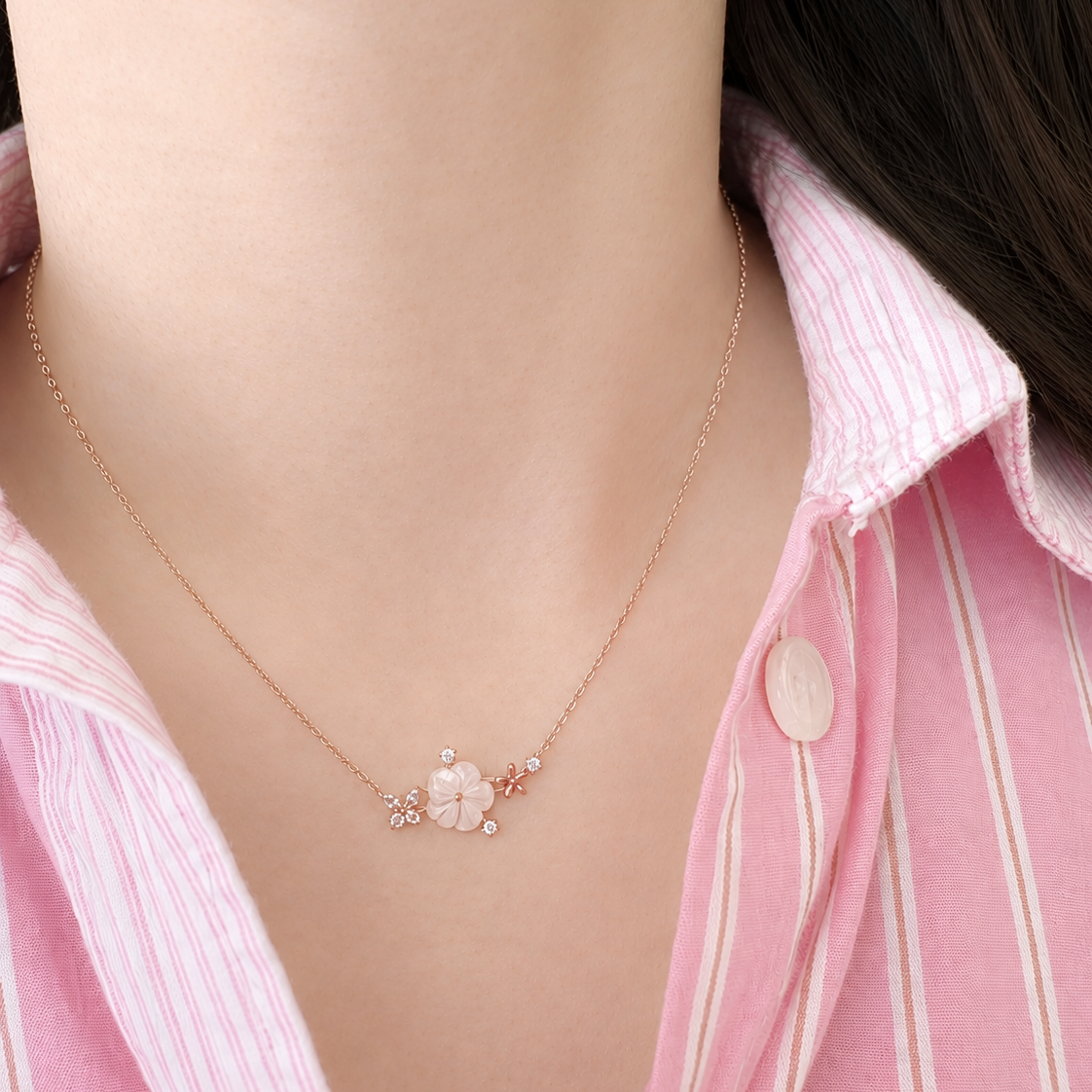 A close-up of a person's neck and chest wearing a delicate rose gold necklace with a small flower and bead pendant, paired with a pink and white striped button-up shirt.
