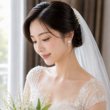 A woman in a white lace wedding dress and veil holding a bouquet of white lily of the valley flowers, photographed indoors near a window.