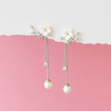 A pair of dangling floral earrings with white flower charms, silver-tone chains, clear rhinestone accents, and pearl-like drops, displayed against a split white and pink background.