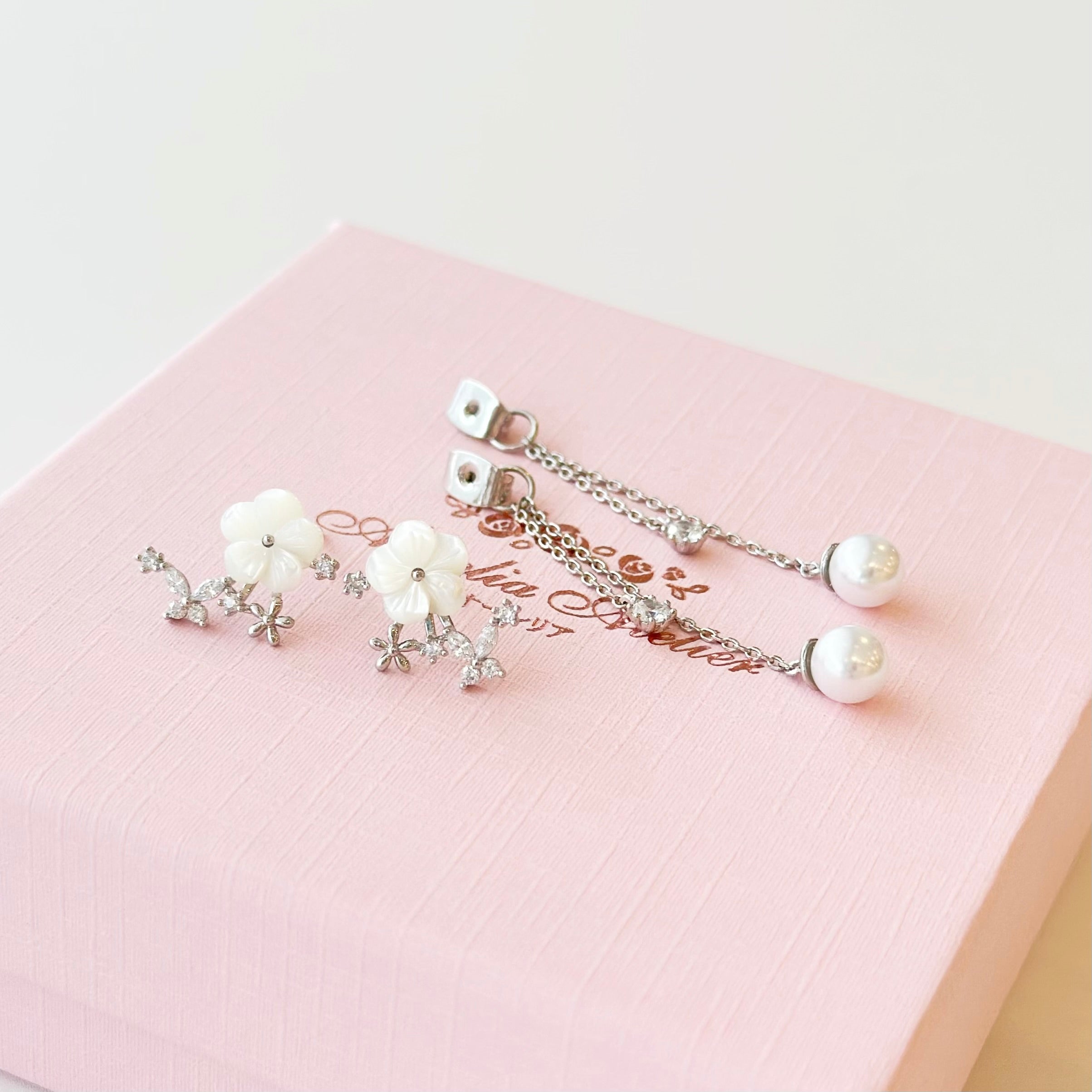 A pair of silver-toned dangle earrings with white flower and pearl details, displayed on a light pink textured jewelry box.