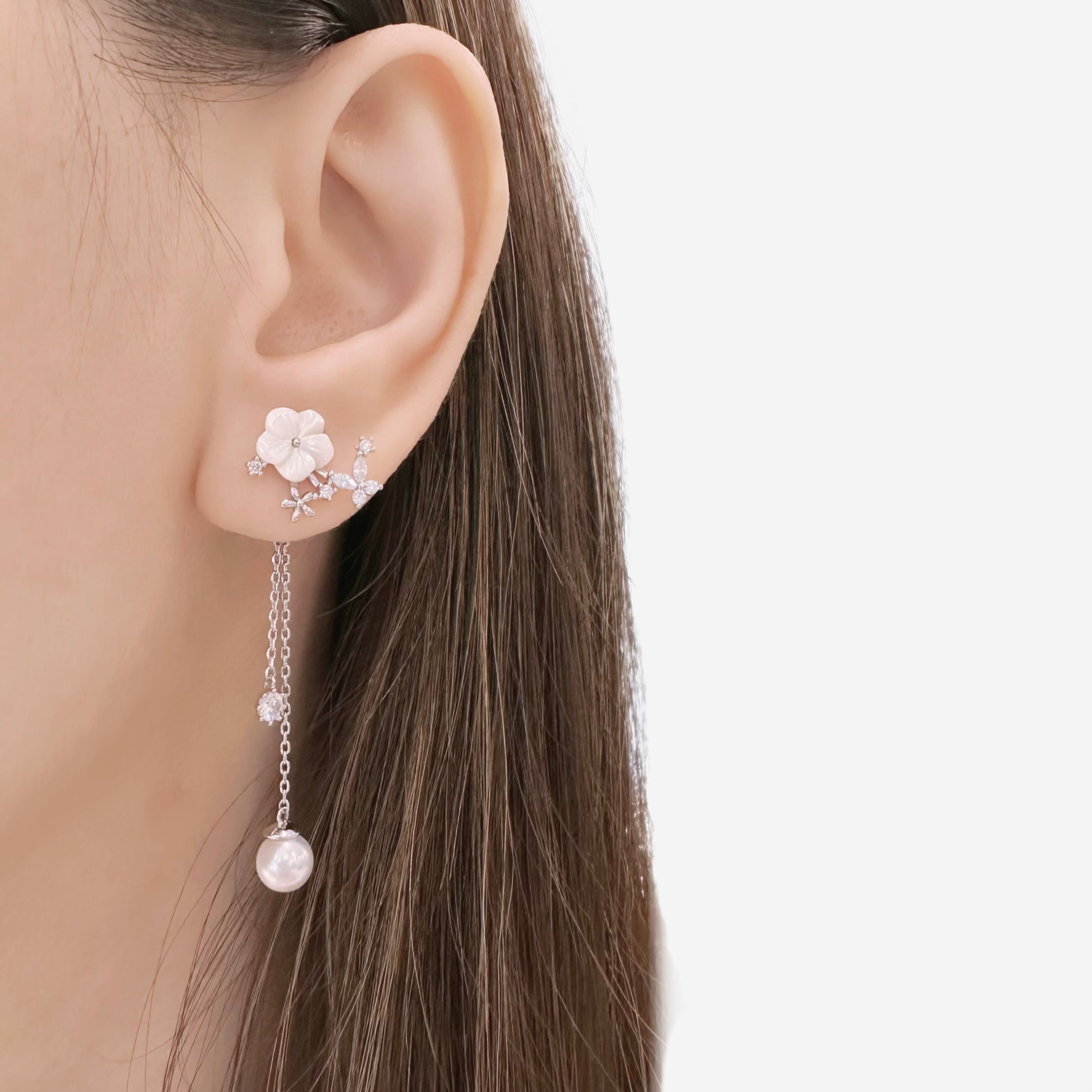 A close-up of a model's ear wearing a silver-toned ear cuff with a white flower and dangling chain with a pearl-like bead, against a light gray background.