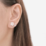 A close-up of a model's ear wearing a delicate floral stud earring in white and silver, with fine leafy sprigs extending from the flower.