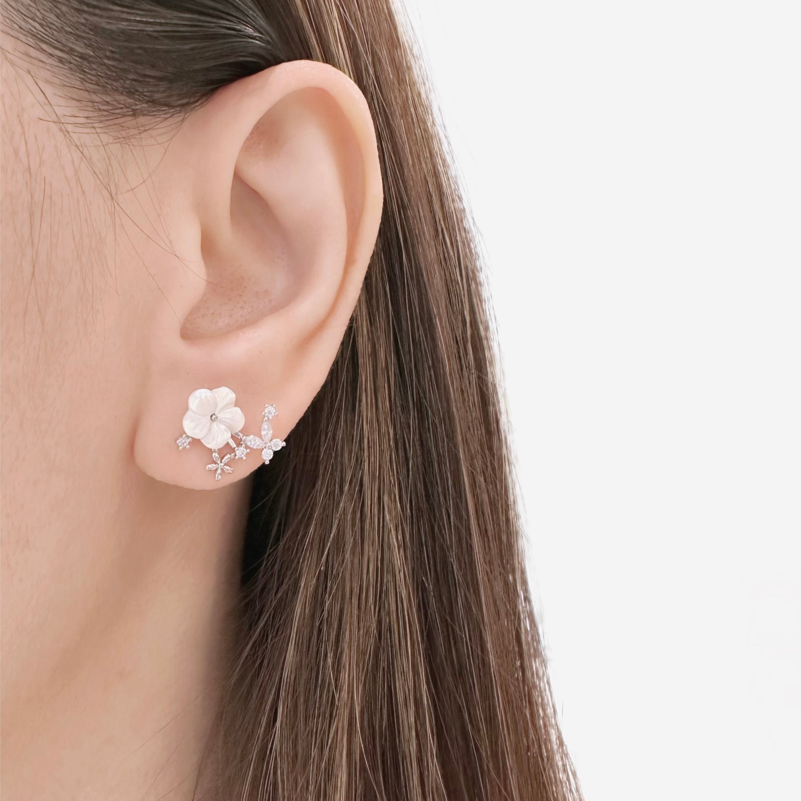 A close-up of a model's ear wearing a delicate floral stud earring in white and silver, with fine leafy sprigs extending from the flower.