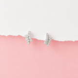 A pair of silver-toned stud earrings with pavé-set clear stones, arranged on a torn-paper style background split horizontally between white on top and pink on bottom.